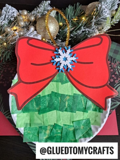 A festive Paper Plate Ornament crafted with green tissue paper squares, a big red paper bow, gold pipe cleaner, and stacked blue and white foam snowflakes. Pine branches and ornaments decorate the background for extra holiday cheer.