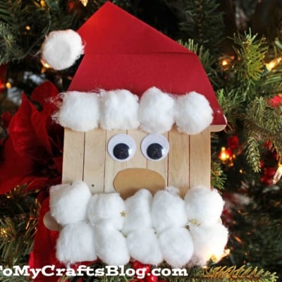 A handmade Popsicle Stick Santa ornament made from popsicle sticks, googly eyes, red paper, and cotton balls hangs on a Christmas tree surrounded by lights and decorations. GluedToMyCraftsBlog.com is at the bottom.