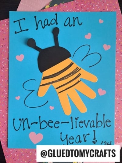A handmade card with a paper bee made from a yellow handprint and black details on blue paper. It says “I had an UN-bee-lievable year!” and makes a charming addition to your collection of new year's keepsakes. Decorated with pink hearts and doodles.