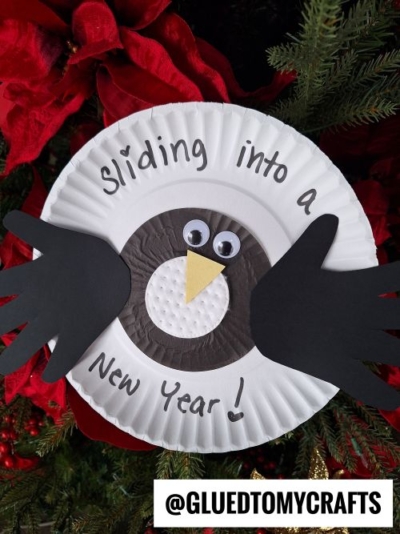 A penguin craft made from a paper plate, black paper wings, googly eyes, and the words Sliding into a New Year!—perfect for new year's keepsakes. Behind it are evergreen branches and red decorations. @GLUEDTOMYCRAFTS is at the bottom.