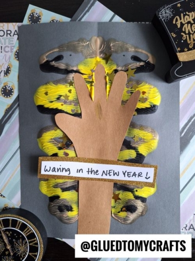 A New Year’s craft featuring a brown paper hand glued on yellow and black painted paper, with a sign reading “Waving in the NEW YEAR!”—a perfect choice for festive new year's keepsakes.