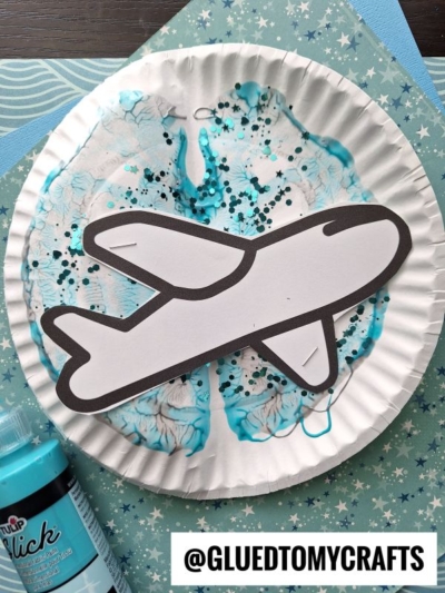 A paper plate airplane craft features a blue, glittery plate as the background for a black-and-white cutout airplane, displayed on star-patterned paper. A bottle of blue paint is nearby—perfect inspiration for how to make a paper plate airplane.