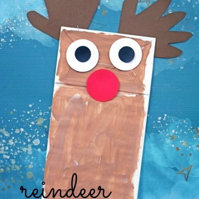 A Paper Bag Reindeer puppet with a brown painted face, large googly eyes, red nose, and brown paper antlers sits on a blue background decorated with gold stars. The text reads reindeer PUPPET.