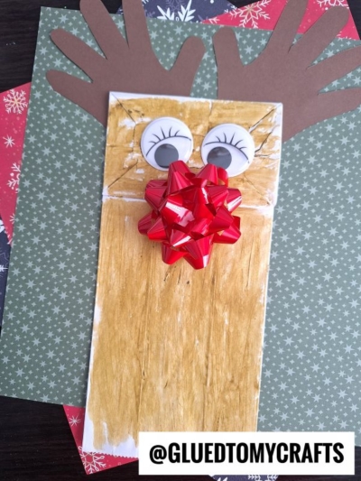 A brown paper bag reindeer puppet decorated as a festive reindeer with googly eyes, handprint antlers, and a red bow for the nose, placed on cheerful paper. The text @GLUETOMYCRAFTS appears at the bottom.