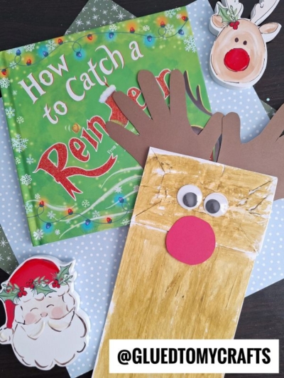A Paper Bag Reindeer puppet with googly eyes and brown handprint antlers sits next to a How to Catch a Reindeer book and cutouts of Santa and a reindeer on a blue polka-dotted surface.