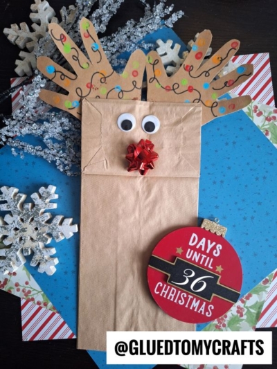 A Paper Bag Reindeer puppet with handprint antlers, googly eyes, and a red bow nose sits on festive paper with snowflake decorations and a “36 days until Christmas” sign. The handle @GLUEDTOMYCRAFTS is displayed.