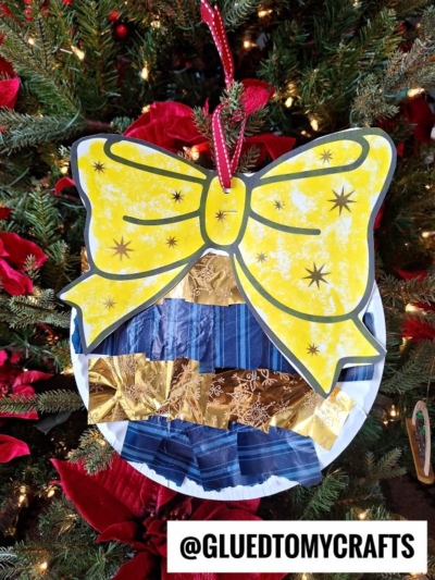 A handmade Paper Plate Ornament craft featuring a large yellow bow and a round shape decorated with blue and gold paper strips, hanging on a Christmas tree with red poinsettias. Text at the bottom reads @GLUEDTOMYCRAFTS.