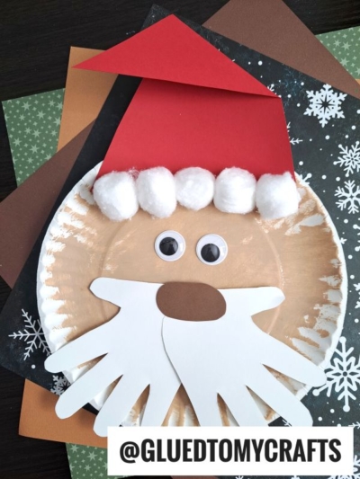 A paper plate Santa craft featuring a red hat, white cotton ball trim, googly eyes, and a handprint beard, displayed on colorful craft paper sheets. The @GLUETOMYCRAFTS handle is at the bottom.