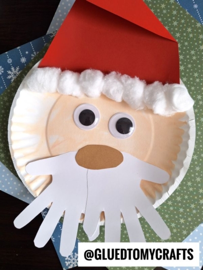 A paper plate Santa craft featuring a handprint beard, cotton ball hat trim, large googly eyes, and a paper nose. Set against blue and green holiday paper, this festive project is perfect for creative holiday fun!.