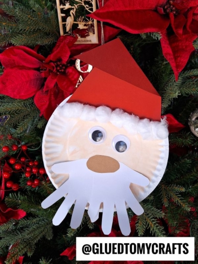 A charming paper plate Santa craft featuring a red hat, cotton brim, googly eyes, and a playful handprint beard, hanging on a Christmas tree with red poinsettias. @GLUEDTOMYCRAFTS is written at the bottom.