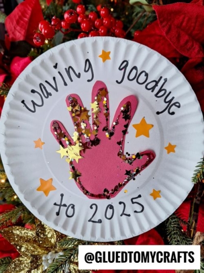 A paper plate with a pink handprint in the center, decorated with gold glitter and star confetti. This charming new year's keepsake says "Waving goodbye to 2025" and is surrounded by festive decorations.
