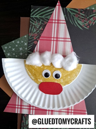 A Paper Plate Santa face craft made from a folded paper plate, with cotton balls for a beard, googly eyes, and a red paper nose, placed on festive patterned paper. @GLUEDTOMYCRAFTS is written at the bottom. Perfect for any paper santa craft project!.