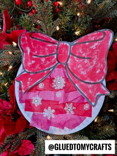A handmade Paper Plate Ornament craft shaped like a round bauble with a large red bow, decorated with tissue paper and snowflake stickers, hangs on a Christmas tree. The handle @gluedtomycrafts appears at the bottom.
