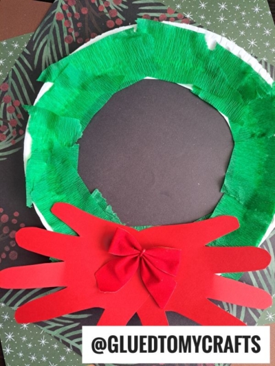 A handmade Paper Plate Christmas Wreath craft made from green tissue paper and red cut-out hand shapes, decorated with a red bow, is displayed on a festive background. The text @GLUEDTOMYCRAFTS is at the bottom.