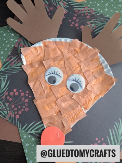 A reindeer face craft made from brown paper squares, large googly eyes, a red paper nose, and handprint antlers—just like a fun paper plate reindeer—on a festive background; @GLUEDTOMYCRAFTS label at the bottom.