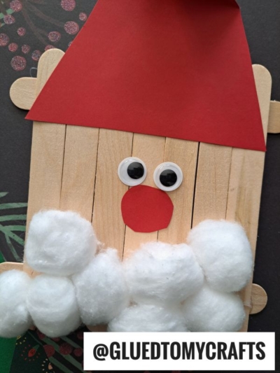 A Popsicle Stick Santa face craft made with popsicle sticks, a red paper hat and nose, googly eyes, and cotton ball beard. Displayed on a dark patterned surface with @GLUEDTOMYCRAFTS at the bottom.