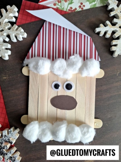 A Popsicle Stick Santa face made with cotton balls for the beard and hat trim, a red-striped paper hat, googly eyes, and a brown paper mouth. Snowflake decorations and a “@GLUEDTOMYCRAFTS” tag are visible.