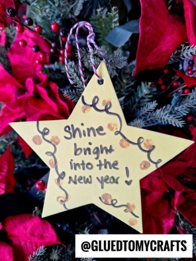 A yellow star-shaped ornament, perfect for new year's keepsakes, with the words Shine bright into the New Year! hangs on a Christmas tree adorned with red flowers and greenery. Confetti-like dots and curly lines decorate the ornament. @GLUEDTOMYCRAFTS is written below.