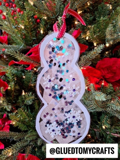A glass snowman ornament with sequins and snowflake confetti hangs on a Christmas tree adorned with red poinsettias and twinkling lights. A red ribbon tops it off. Text at the bottom reads @GLUEDTOMYCRAFTS.