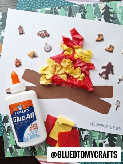 A paper campfire craft made from brown paper logs and crumpled red and yellow tissue paper for flames. Elmer’s Glue and spare tissue paper are nearby, with camping-themed stickers decorating the background.