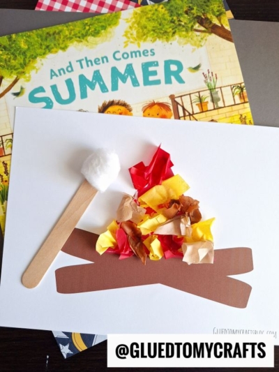 A children’s summer craft features a paper campfire craft made of brown paper logs and red, orange, and yellow tissue “flames” next to a craft stick marshmallow, all resting atop a summer-themed picture book.