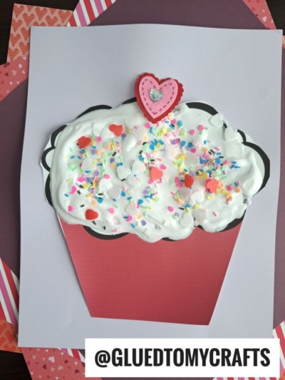 A handmade Puffy Paint Cupcake craft with a red wrapper, white textured topping, colorful sprinkles, and a layered pink and red heart on top, displayed on a white sheet with a patterned background. @GLUEDTOMYCRAFTS is written below.