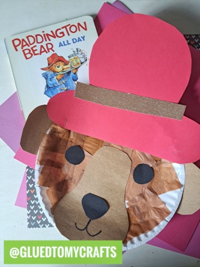 A Paddington Bear book sits next to a charming paddington bear paper plate craft, complete with brown paper fur and a large red hat, all arranged on pink and patterned paper. A green label reads @GLUEDTOMYCRAFTS.