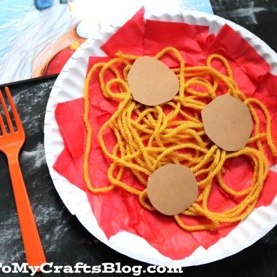 A paper plate displays yellow yarn as paper spaghetti and three brown paper circles resembling meatballs, with red tissue paper sauce underneath. An orange plastic fork sits beside the Paper Plate Spaghetti craft. GluedToMyCraftsBlog.com is at the bottom.