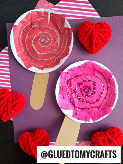 Two diy paper plate lollipops decorated with red and pink tissue paper and glitter spirals are displayed on a purple background, surrounded by yarn-wrapped red hearts and striped paper accents.