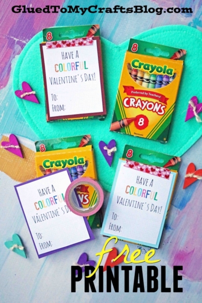 Flat lay of two boxes of Crayola crayons, Valentine’s cards reading "Have a Colorful Valentine's Day! To: From:", a roll of washi tape, heart cutouts, and free printable text set on a blue heart background.