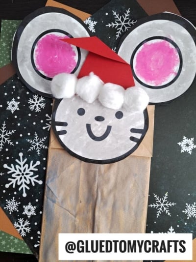 A paper bag mouse craft featuring a red Santa hat with cotton balls, large pink ears, and a smiling face, placed on snowflake-patterned and brown paper. Try using a mouse paper bag puppet template! @GLUEDTOMYCRAFTS is written at the bottom.