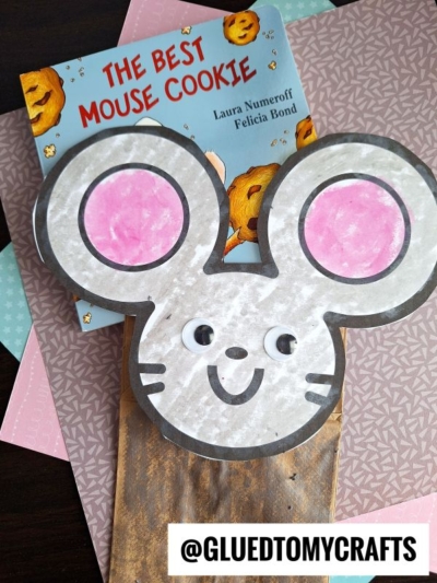A children’s book titled The Best Mouse Cookie sits on colored paper, partially covered by a mouse paper bag puppet with big pink ears and googly eyes. A @GLUEDTOMYCRAFTS label is at the bottom.