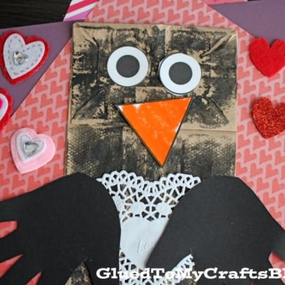 A paper bag penguin with big googly eyes, an orange beak, hand-shaped wings, a white doily belly, and heart shapes sits on a purple background.