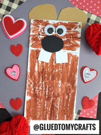 A paper bag craft painted brown to resemble a beaver or Paper Bag Groundhog, with large teeth, round black nose, and tan ears, surrounded by red and pink felt hearts on a colorful background.
