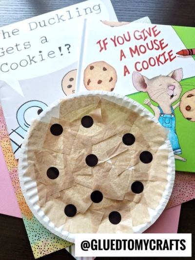 A paper plate cookie kid craft resembling a chocolate chip cookie is shown with children's books If You Give a Mouse a Cookie and The Duckling Gets a Cookie!? in the background, along with colorful papers.