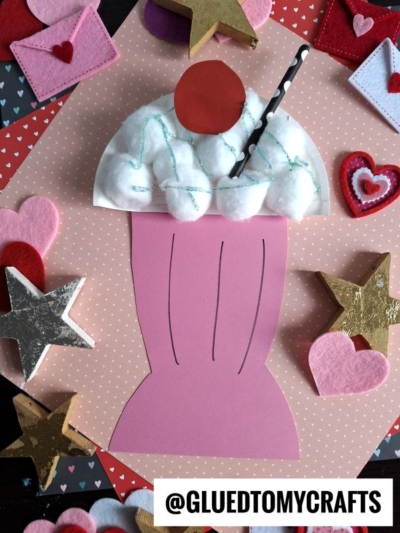 A Paper Plate Milkshake craft features a pink glass, cotton ball whipped cream, a red cherry, and a striped straw on a pink polka dot background with felt hearts, stars, and mini envelopes. @GluedToMyCrafts is written at the bottom.
