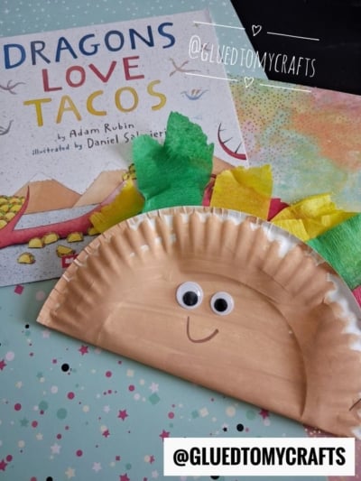 A cheerful paper plate taco craft with googly eyes and a smile sits on top of the book Dragons Love Tacos. Colorful tissue paper lettuce and @GLUEDTOMYCRAFTS are also visible in the image, making this Paper Plate Taco project both playful and fun.