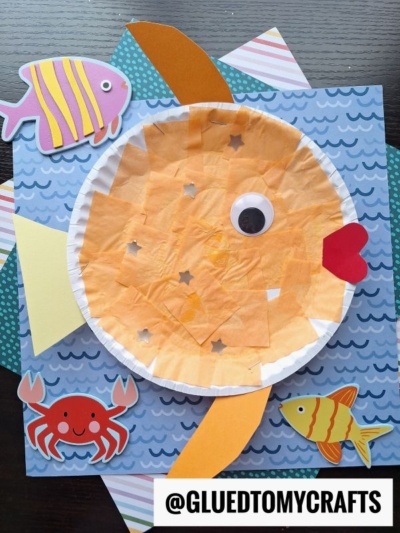 A paper plate goldfish craft with orange tissue paper, a googly eye, and red heart-shaped mouth sits among paper cutouts of a Dr Seuss fish, yellow fish, and red crab on a blue wavy background. @GLUEDTOMYCRAFTS is written at the bottom.