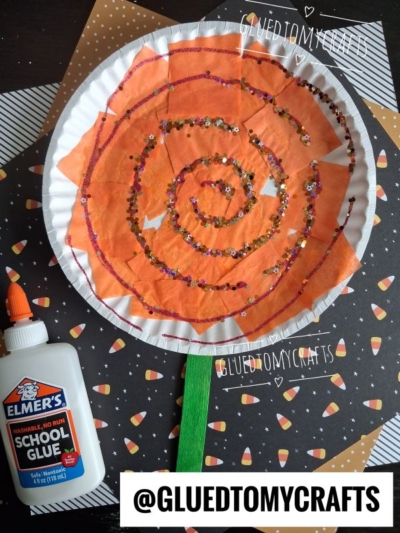 A large Paper Plate Lollipop craft with an orange swirl and glitter shines brightly, featuring a green stick handle and a school glue bottle nearby, all set on a candy corn-themed background. If you’re looking for inspiration on how to make paper plate lollipops, this eye-catching creation is sure to spark creativity—@GLUEDTOMYCRAFTS is written at the bottom.