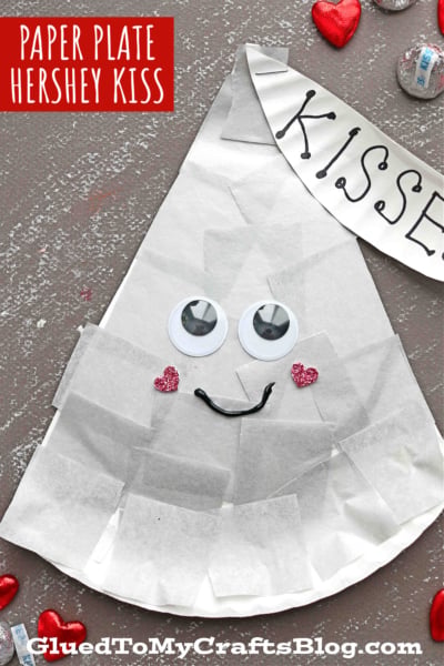 A Paper Plate Hershey Kiss decorated with white tissue paper, large googly eyes, pink glitter heart cheeks, a smiling mouth, and a white KISSES banner sits on a gray surface with red hearts and Hershey’s Kisses.