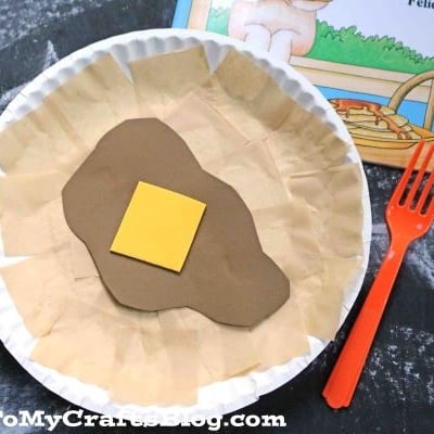 A Paper Plate Pancake craft made with beige and brown paper and topped with a yellow butter square sits next to a red plastic fork and a partially visible children's book.