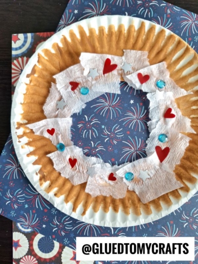 A paper plate doughnut with a brown rim is decorated with white crepe paper, red heart shapes, and blue gems, placed on dark blue firework-patterned paper. A label reads @GLUEDTOMYCRAFTS—a sweet idea for a donut kid craft!.
