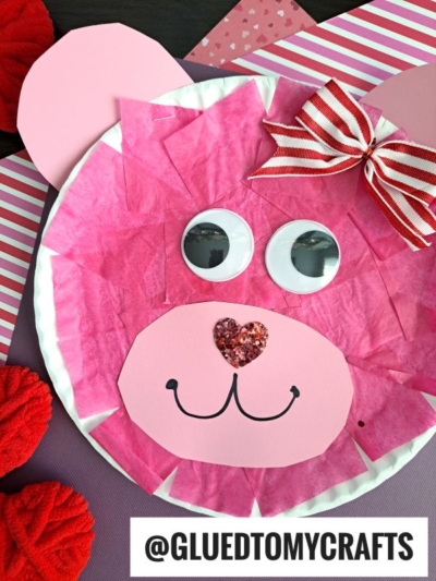 A pink paper plate bear with tissue paper fur, googly eyes, a red glitter heart nose, and a bow smiles brightly—this valentine bear craft is perfect as a cute bear craft for kids on colorful striped and solid backgrounds.