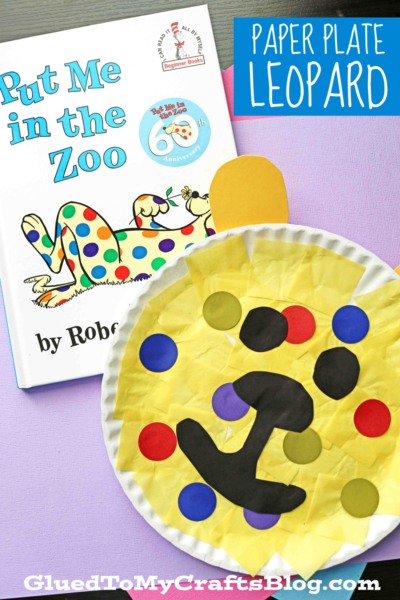A children’s book titled Put Me in the Zoo lies next to a colorful paper plate leopard craft, decorated with bright colored paper spots and yellow tissue paper. The scene is set against a light lavender background.