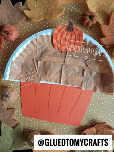 A Paper Plate Muffin-inspired cupcake craft made with brown paper, a paper pumpkin on top, and an orange paper base, surrounded by felt leaves and small pumpkins on a polka dot background. Text reads @GLUEDTOMYCRAFTS.