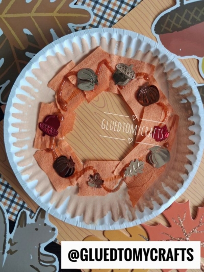 A paper plate donut decorated with orange paint, tissue paper pieces, and autumn-themed buttons shaped like pumpkins and leaves. Background includes fall-themed patterns and the text @GLUEDTOMYCRAFTS, making this doughnut kid craft a perfect seasonal project.
