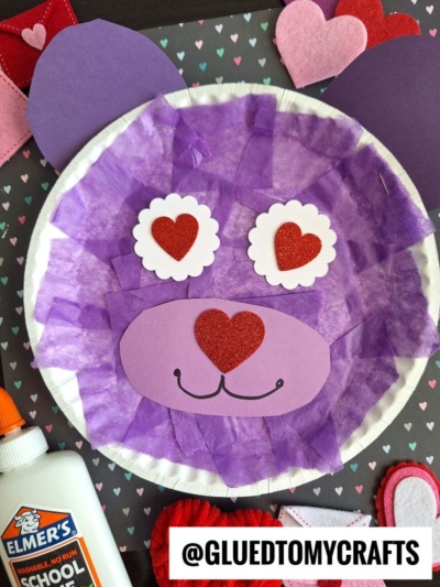 A cute paper plate bear craft made with purple tissue paper, heart-shaped nose, and heart eyes, perfect as a valentine bear craft for kids. Surrounded by felt hearts, Elmer’s glue, and a @GLUEDTOMYCRAFTS label at the bottom.