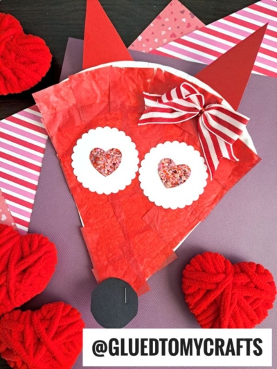 A Paper Plate Valentine Fox craft featuring a red fox face made from tissue paper, paper shapes, and a red ribbon bow, with heart-shaped eyes, displayed on pink and purple paper and surrounded by red yarn hearts.