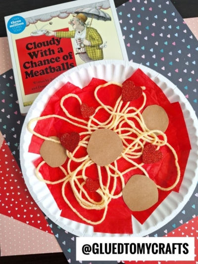 Paper Plate Spaghetti craft featuring yellow yarn as paper spaghetti, red tissue as sauce, brown paper for meatballs, and glitter hearts. The book Cloudy With a Chance of Meatballs is in the background on heart-patterned paper.