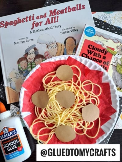 A Paper Plate Spaghetti craft features yarn “spaghetti” and brown paper “meatballs” on a red and white plate, alongside books like “Spaghetti and Meatballs for All!,” Elmer’s glue, and crafting supplies. @gluedtomycrafts.